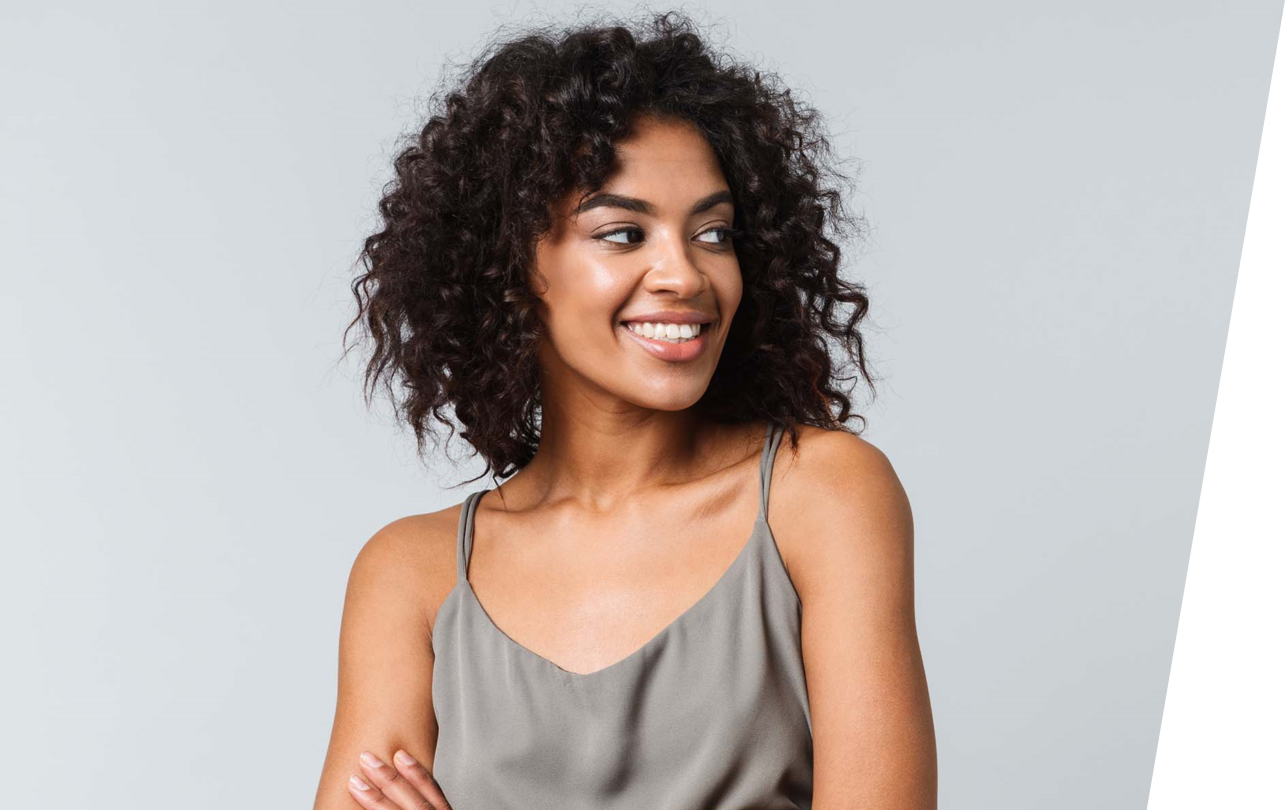 Woman with natural curls and effortless style stands, arms folded, in a silver satin tank, looking away from camera, smiling.
